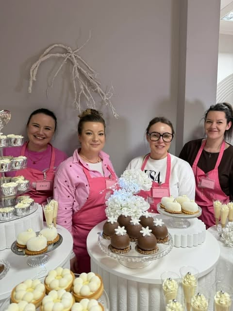 Four women in pink aprons stand behind a display table of cupcakes, cakes, and desserts at a bakery event