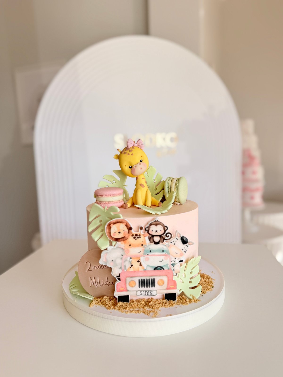 Whimsical tiered cake featuring safari animals, giraffe topper, pink jeep toy, and green leaf decorations under a dome