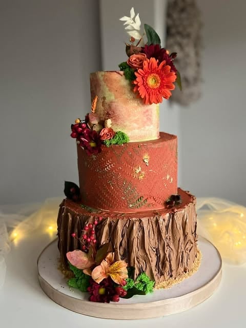Four-tiered decorative cake with coral, rose gold, and brown textures, adorned with red and coral flowers and greenery