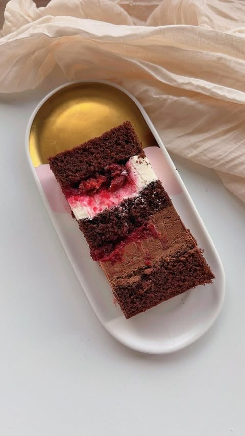 Layered chocolate cake slice with pink frosting and gold sauce on white plate with beige fabric background