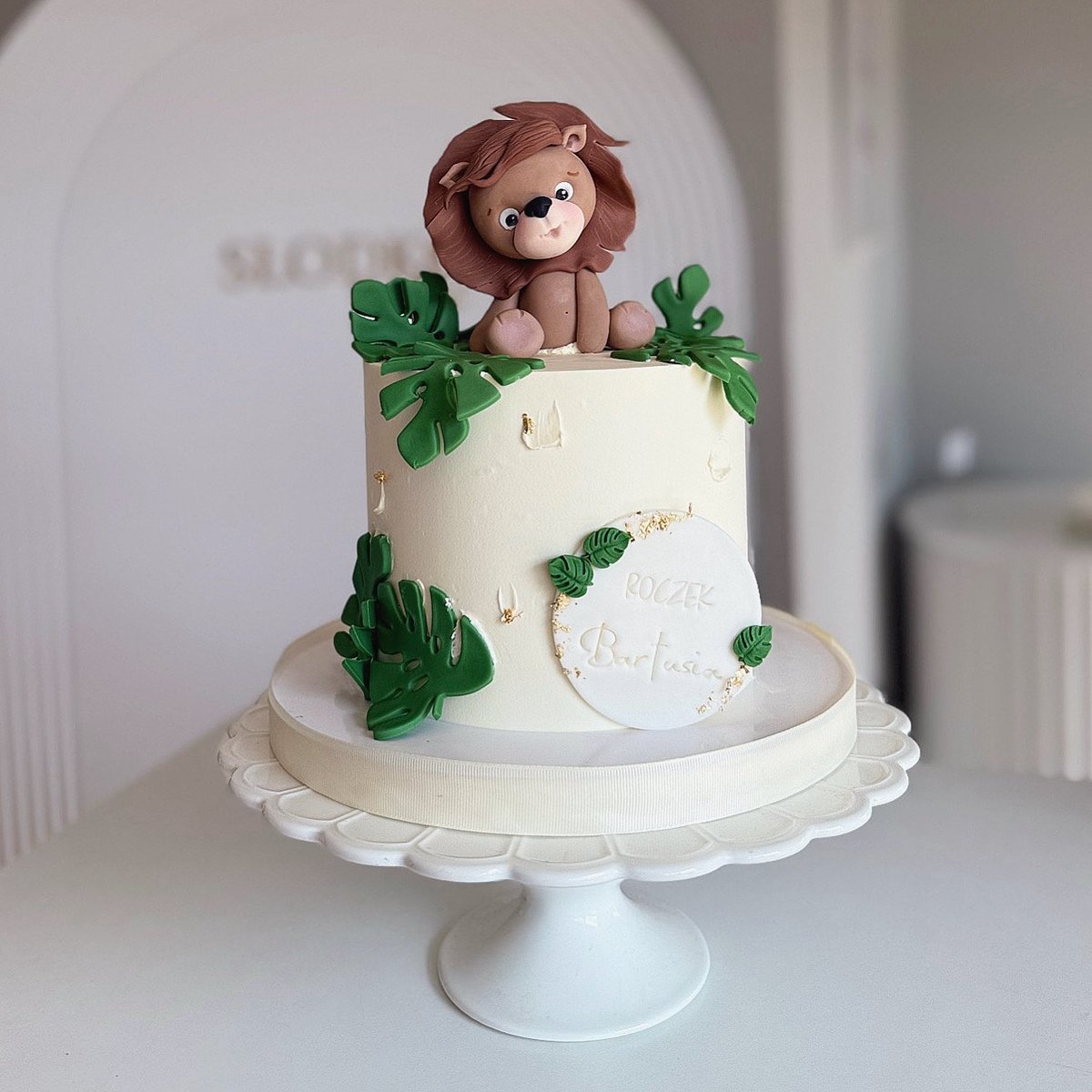 Two-tiered white cake with brown fondant lion on top and green sugar leaves on white cake stand