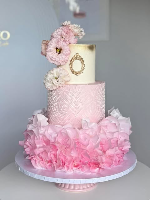 A three-tiered elegant pink and gold wedding cake decorated with peonies, lace texture, ruffled fondant, and an ornate gold frame detail