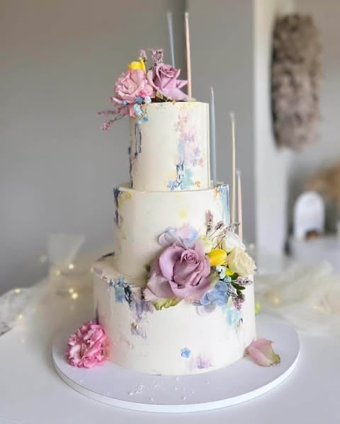 Three-tiered white wedding cake decorated with pink and purple flowers, yellow accents, and tall silver candles on a white plate
