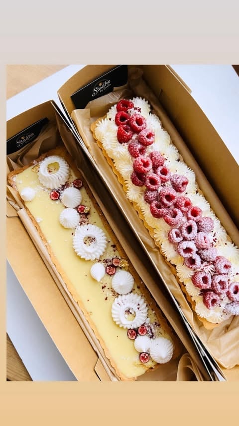 Two elegant pastry desserts in cardboard boxes: one with white cream and red berries, one with yellow custard and white meringues
