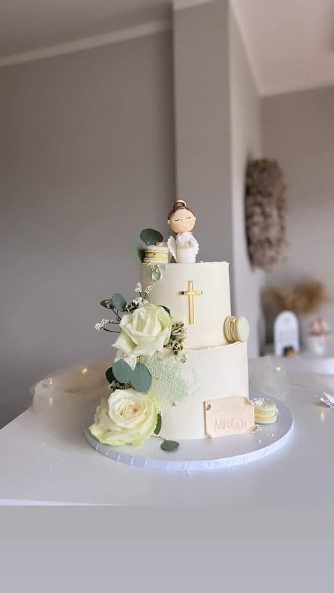 Multi-tiered white wedding cake with gold cross, cream roses, greenery, figurine topper, and macarons on a white cake stand