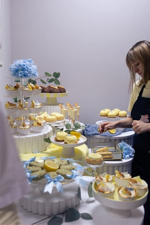 Woman arranging desserts and pastries on tiered white cake stands decorated with blue flowers and yellow accents at an elegant event display