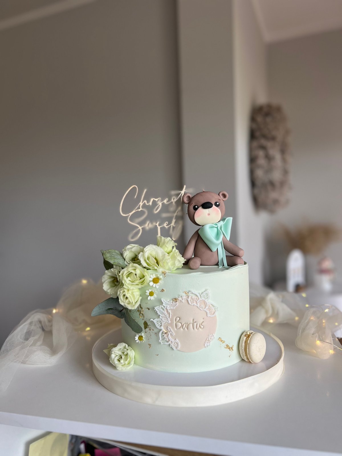 Elegant baby shower cake with pale green frosting, white flowers, teddy bear topper, and Chosen Speci script topper on white cake stand