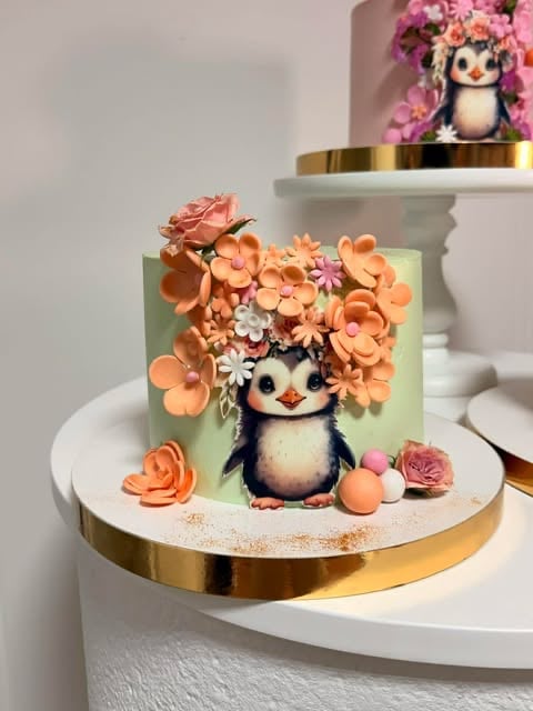 Decorated cake with adorable penguin character and peach-colored sugar flowers on sage green background with gold trim