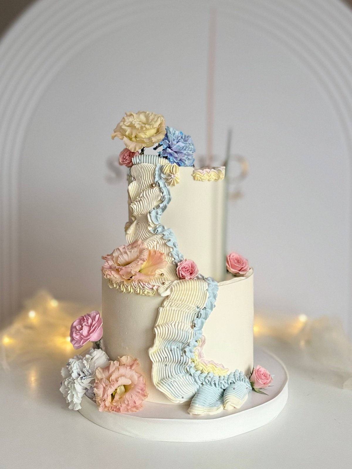 Two-tier white wedding cake decorated with colorful sugar flowers and delicate ruffled details, displayed on a white surface with warm lighting
