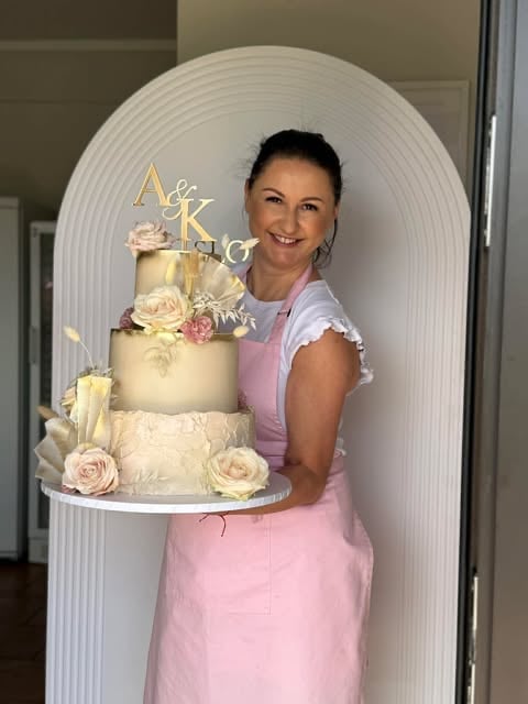Baker holding a three-tier wedding cake with pink roses and gold topper in front of a white arch backdrop