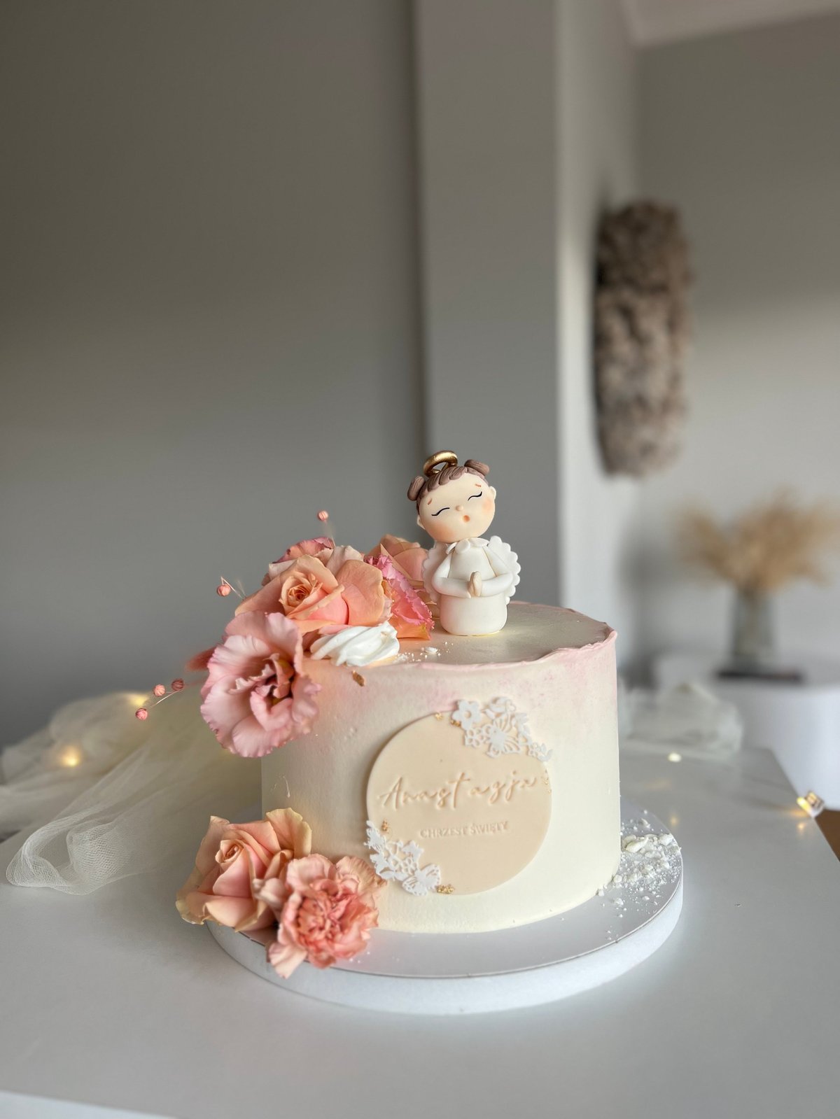 Elegant white cake with pink flowers and angel figurine topper on white plate with fairy lights