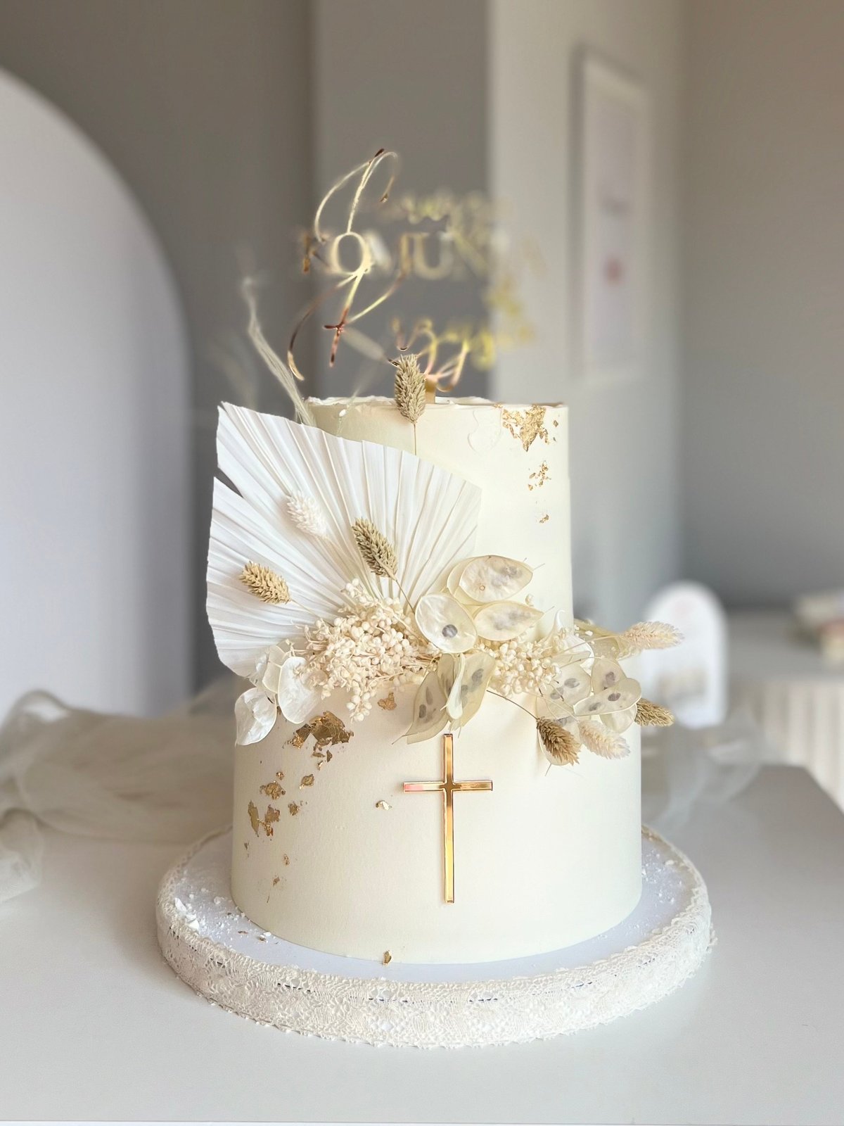 Elegant cream and gold tiered cake decorated with white feathers, dried flowers, a gold cross, and gold branches on a sparkly base