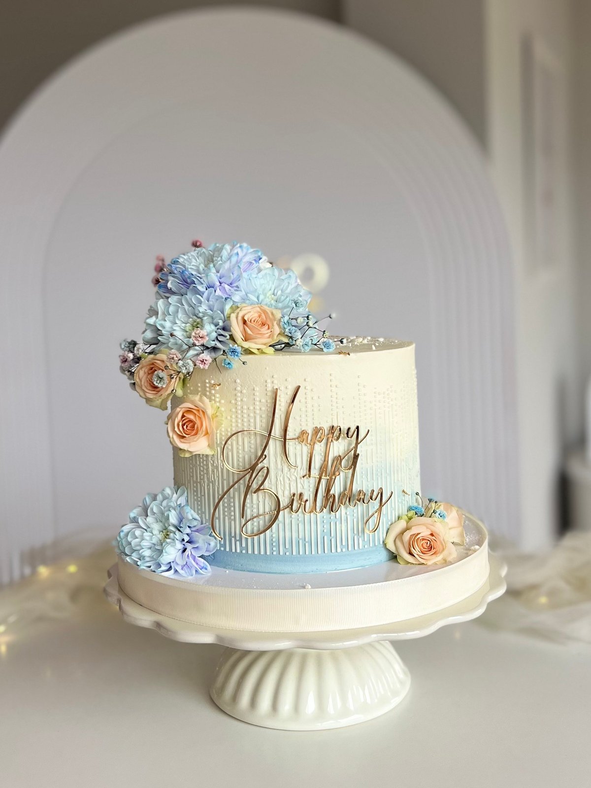 Two-tier birthday cake with blue hydrangeas, peach roses, and gold script on a white cake stand with textured details