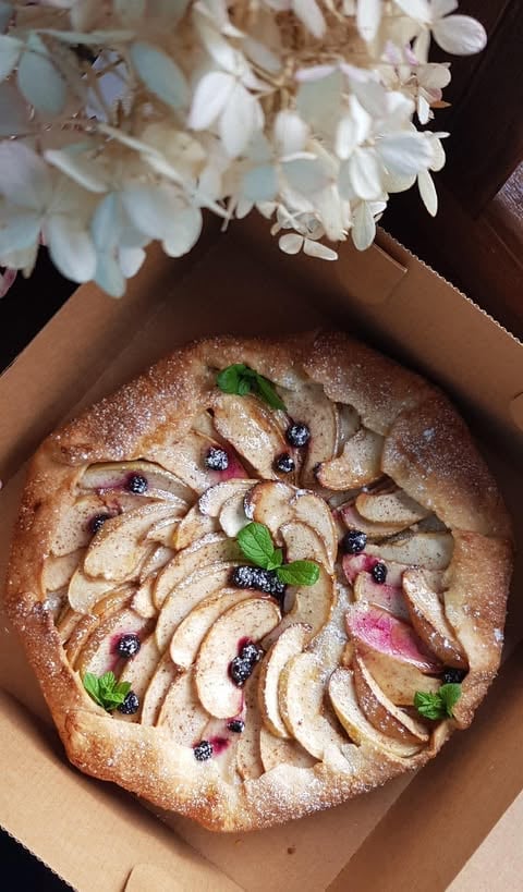 Artisan cake with sliced apples, berries, and fresh mint garnish, dusted with powdered sugar in cardboard box
