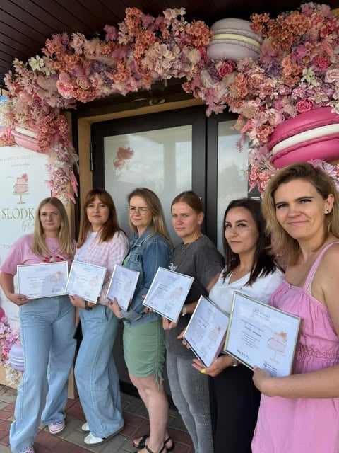 Six women holding certificates standing in front of a floral pink flower installation and storefront