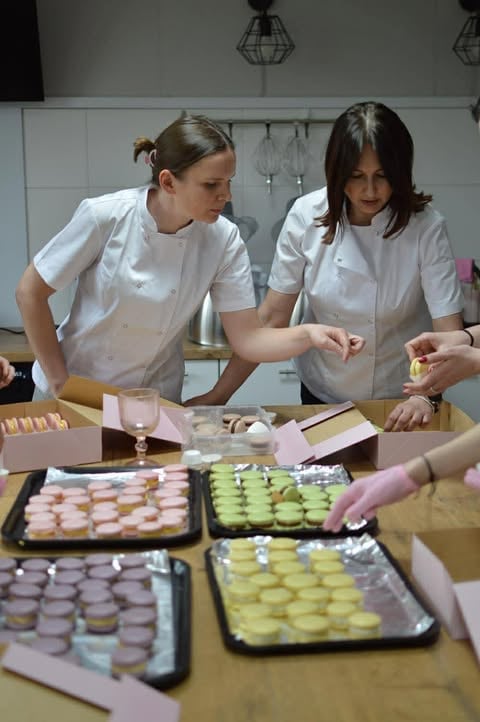 Two women in white chef coats working at a pastry table with multiple trays of decorated macarons and small cakes