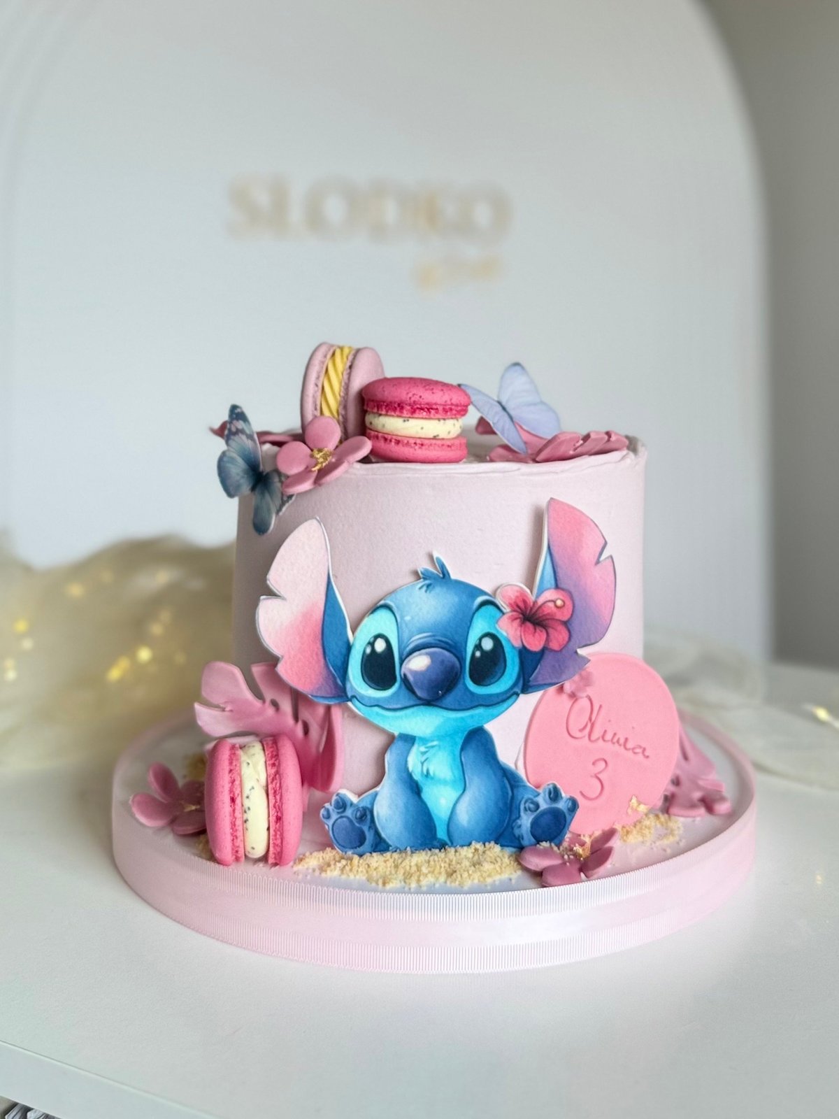 Pink tiered cake decorated with blue Stitch character figurine, macarons, flowers, and gold accents on a white plate