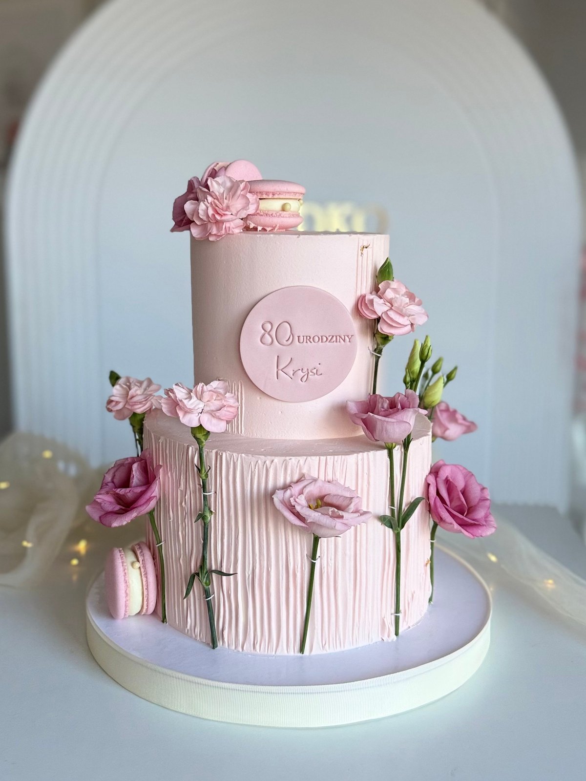 Two-tiered pink birthday cake decorated with fresh carnations and featuring 80 bronzed text on the middle tier, displayed on a white pedestal