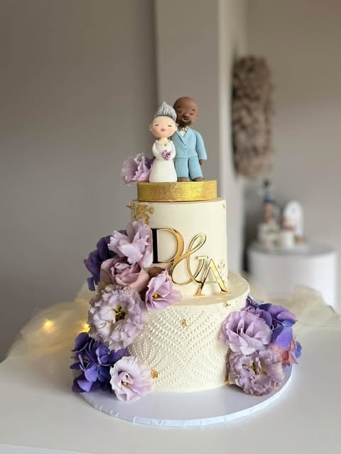 Three-tier wedding cake with white and cream fondant, purple and lavender flowers, golden accents, and bride and groom figurines on top