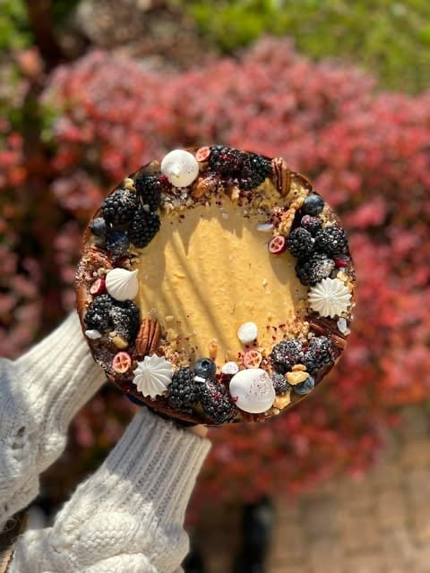 Hand holding a decorated round dessert with gold filling, black berries, and pearl decorations, with blurred pink flowers in background