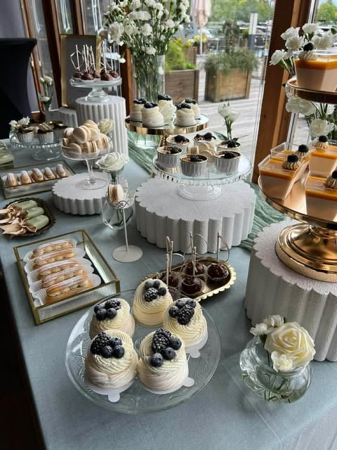 Elegant dessert display with individual cream cakes topped with berries, pastries, and white flowers on tiered stands by bright windows