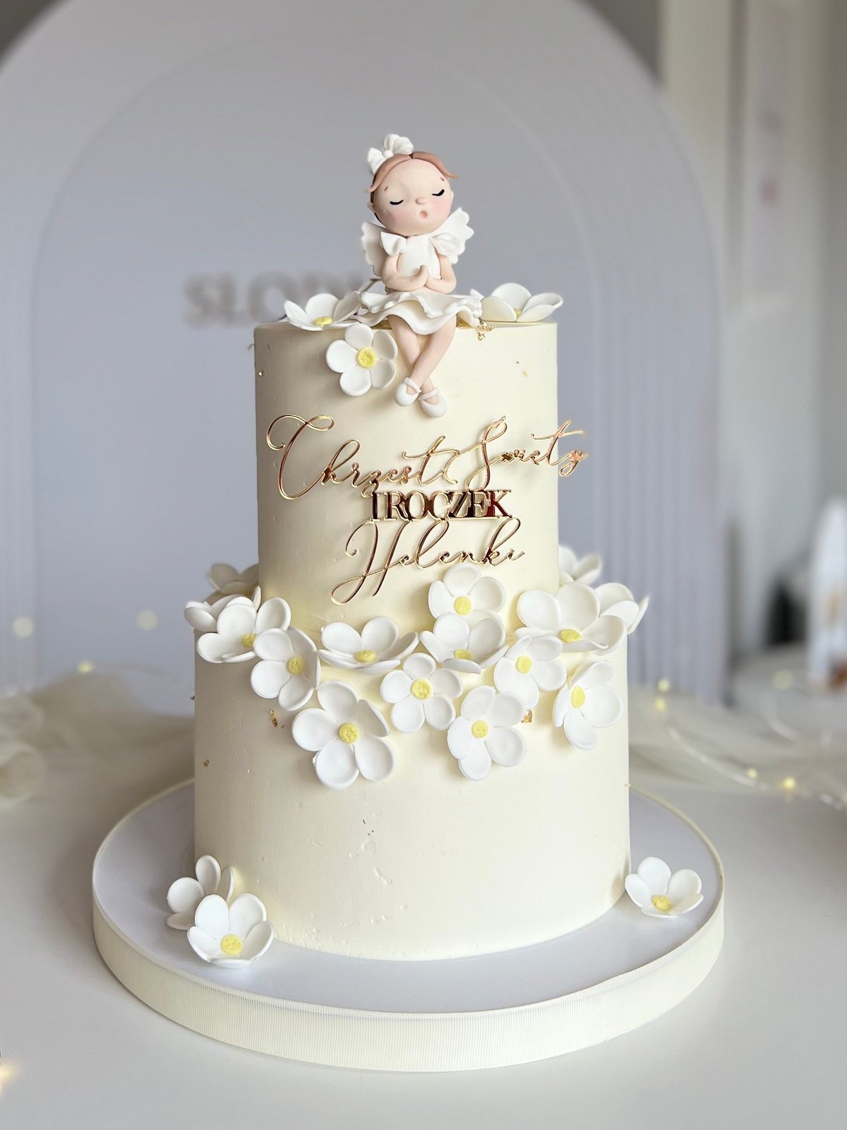 Three-tier cream wedding cake decorated with white fondant flowers and a baby figurine topper