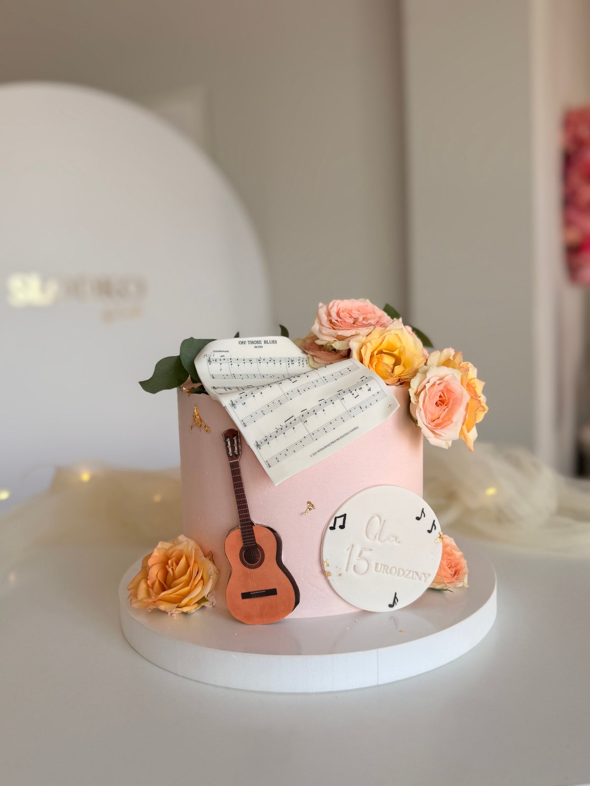 Decorated birthday cake with pink fondant, miniature guitar, sheet music, and fresh orange and pink roses on white base