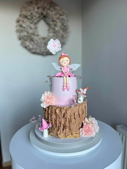 Two-tier fairy themed cake with pink frosting, wood bark texture, fondant fairy figurine, and floral decorations on a blue cake stand