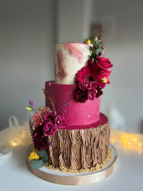 Four-tier decorative cake with textured base, pink and white upper tiers, adorned with burgundy flowers and gold accents