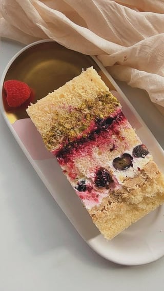 Slice of layered cake with pink frosting, berries, and pistachio crumble on a white plate