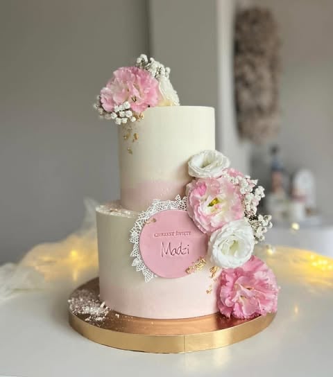 Elegant three-tiered white and pink wedding cake on gold base with fresh pink and white flowers, personalized disc, and sparkly details