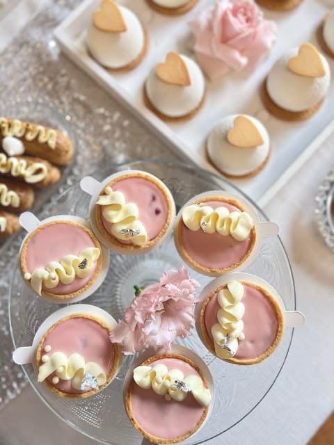 Gourmet dessert display featuring individual tarts with pink glaze and gold piped details, alongside cream-topped cookies with heart decorations