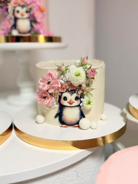 Decorated cake with penguin illustration and fresh pink and white flowers on gold-rimmed base with matching penguin decoration in background