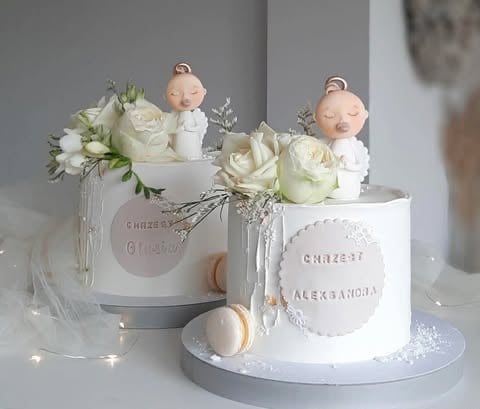 Two elegant white cakes decorated with fresh white roses, baby doll toppers, and delicate flowers in glass vases on white pedestals