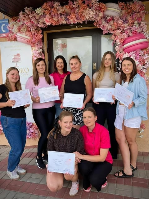 Group of eight women holding certificates standing in front of pink floral decoration and doorway