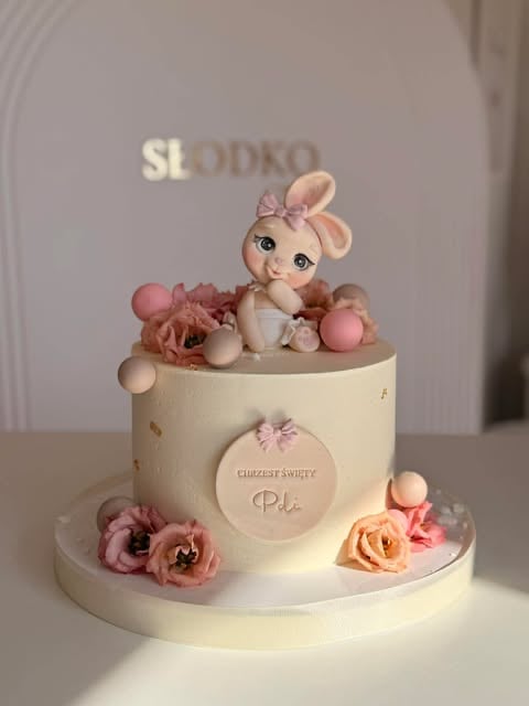 Elegant two-tier white cake with fondant bunny topper, pink flowers, and decorative macarons