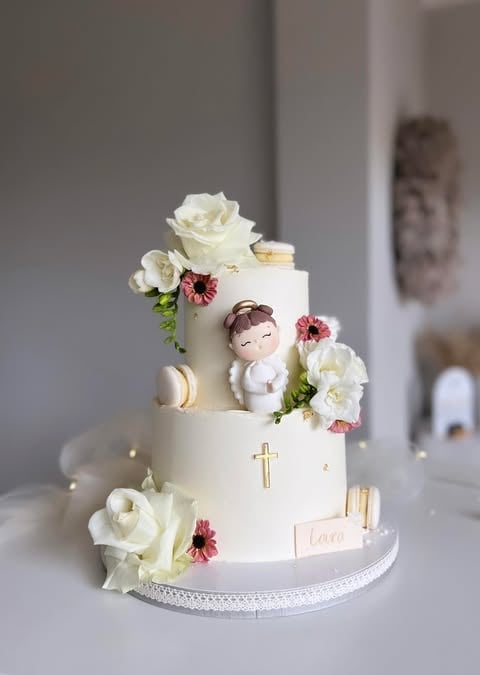 Multi-tiered white baptism cake decorated with cream roses, pink gerbera daisies, a cherub figurine, gold cross, and gold accents on a lace-trimmed cake stand