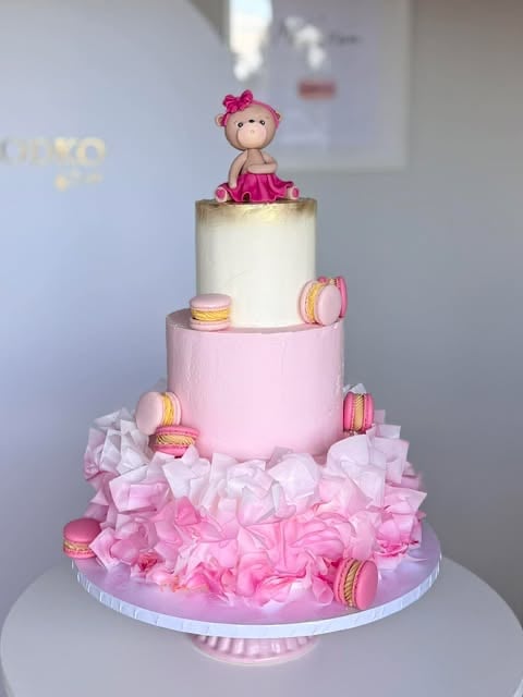 Three-tiered pink and white cake decorated with macarons and ruffled fondant, topped with a doll figure in pink dress