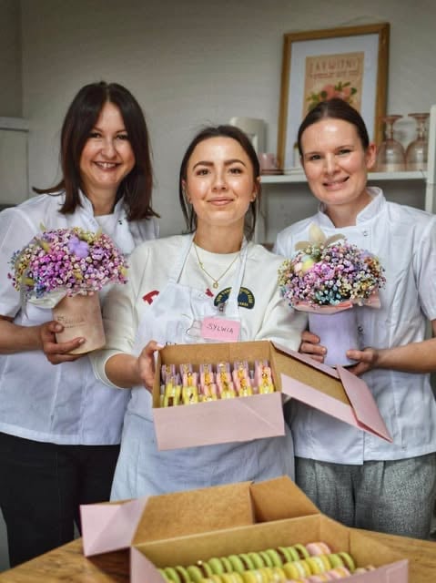 Three women in white uniforms holding flower arrangements and gift boxes in a florist shop