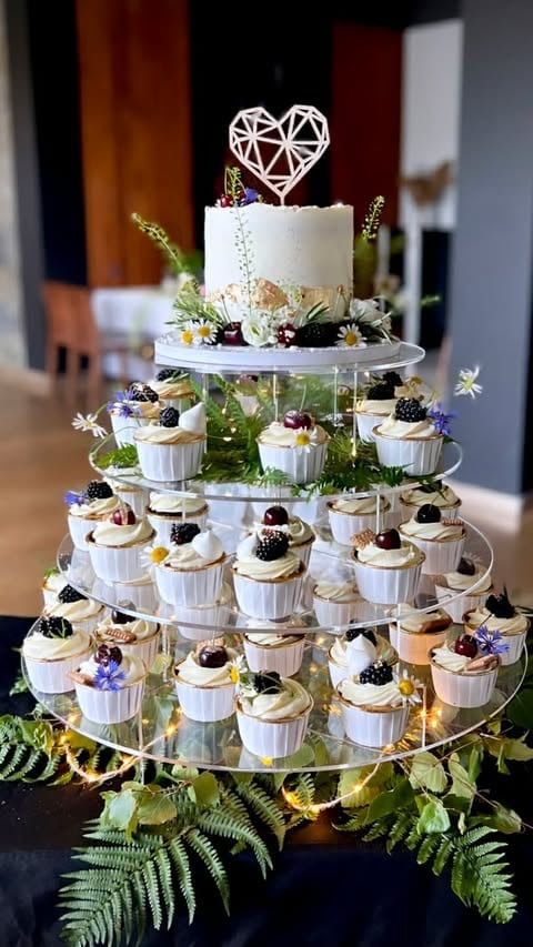 Multi-tiered wedding cake and cupcake tower with white frosting, berries, flowers, and geometric heart topper