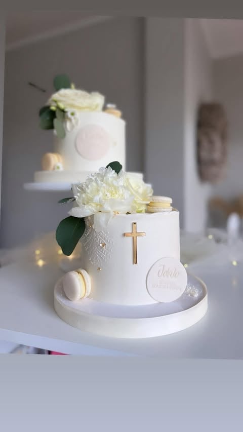 Two-tiered white christening cake with cream flowers, gold cross, green foliage, and macarons on white cake stand