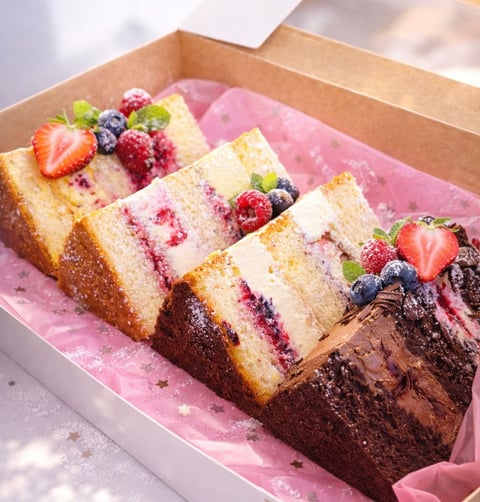Assorted cake slices in a cardboard box lined with pink paper, topped with fresh berries and powdered sugar