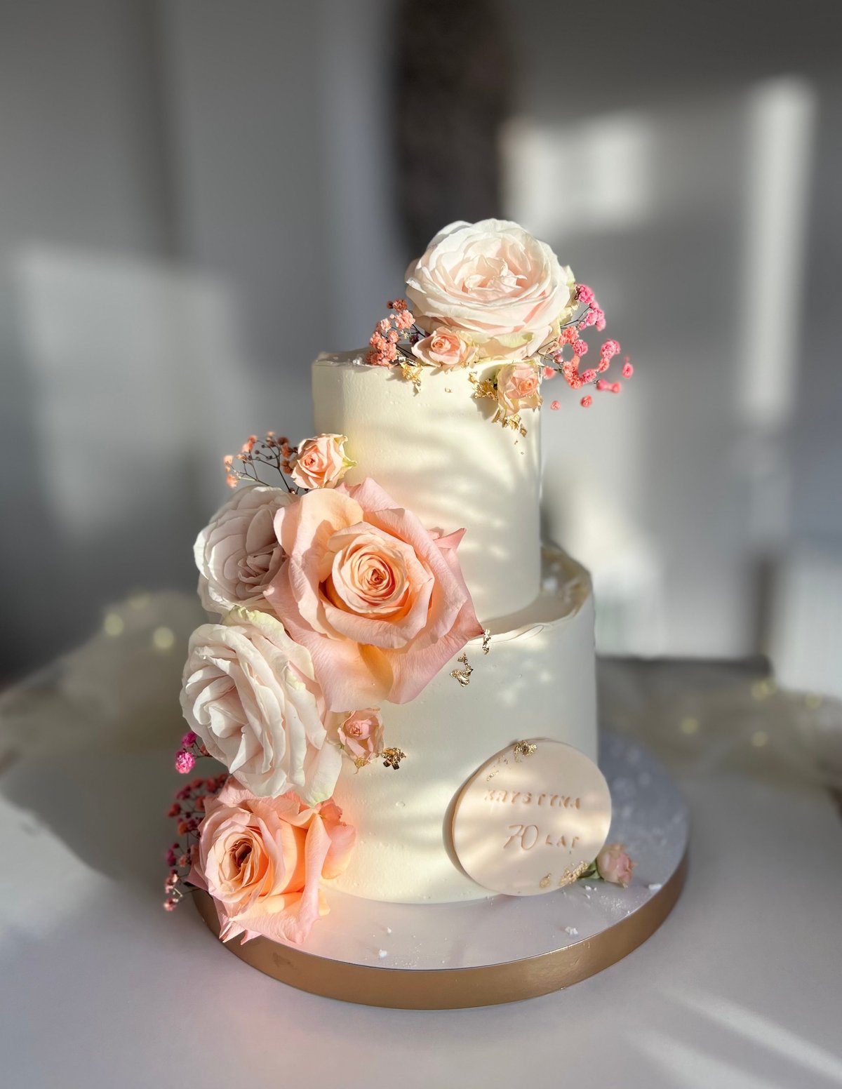 Three-tiered white wedding cake decorated with fresh peach and cream roses, small pink berries, and a personalized cake topper on a gold base