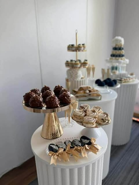 Elegant dessert display with tiered white pedestals featuring chocolate truffles, cupcakes, macarons, and decorative topper cakes on gold accents