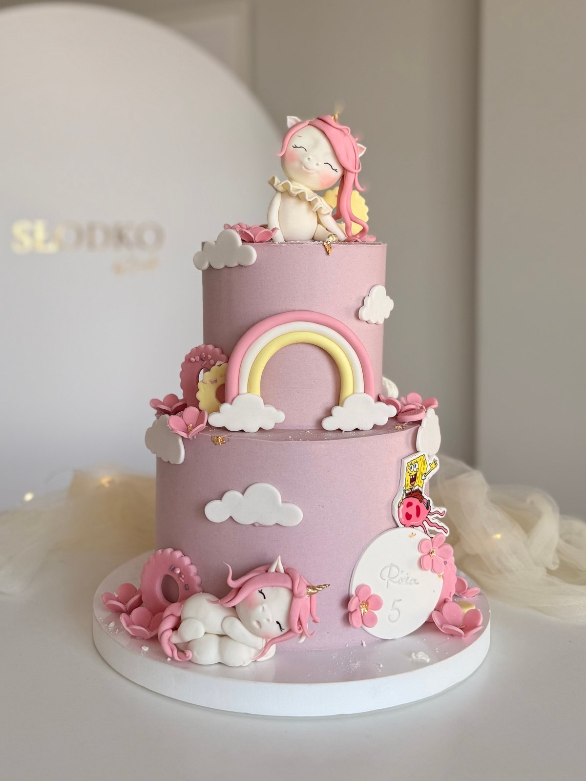 Two-tiered pink fondant cake decorated with pastel clouds, a rainbow, cartoon character figurine on top, and pink floral details on a white base