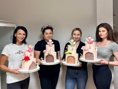 Four women holding decorative tiered cakes with pink flowers and castle-themed designs at an indoor event