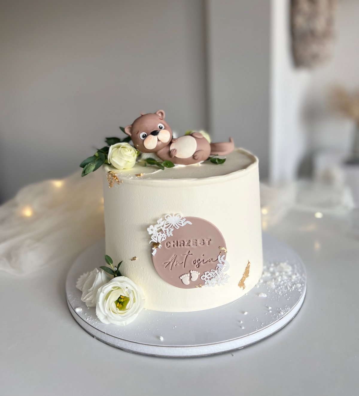 Elegant white fondant cake with decorative bear figurine, flowers, and rose gold accents on a gray plate