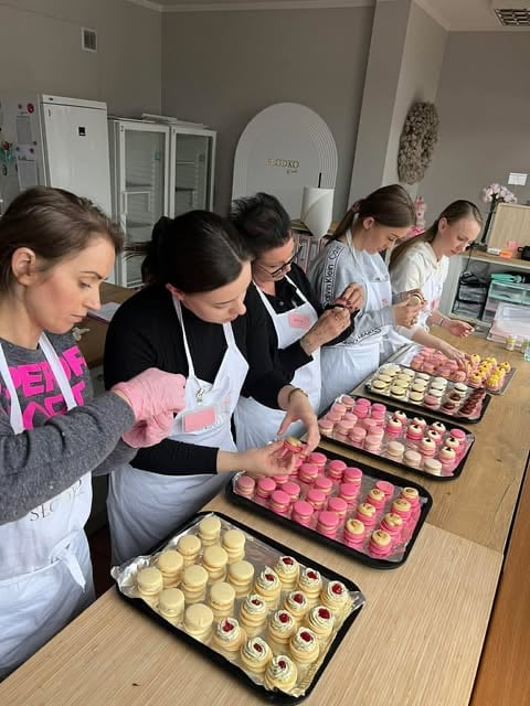 Five people in aprons working on cupcakes arranged in trays on a wooden table in a kitchen
