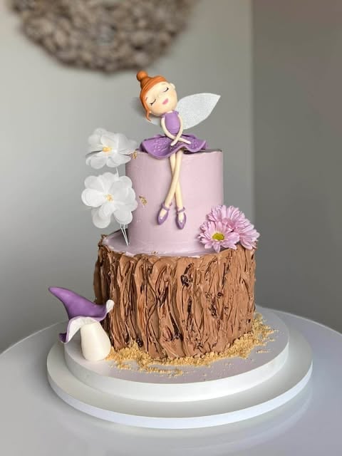 A tiered fairy-themed cake with a fairy figurine on top, featuring white flowers, textured bark design, and purple accents on a white stand