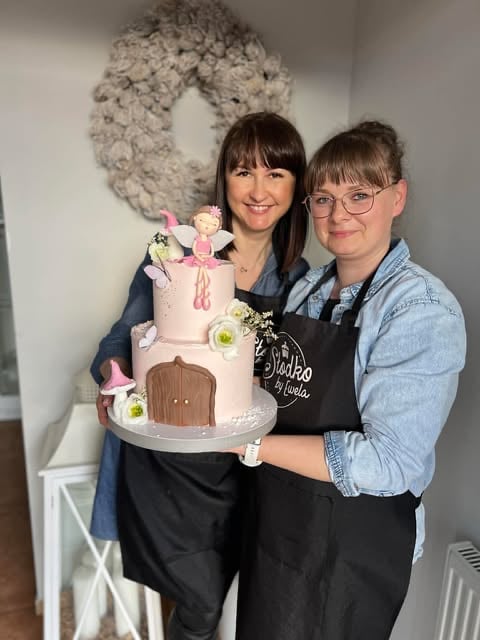 Two women displaying a decorative tiered cake with fairy tale design, standing in front of a wreath